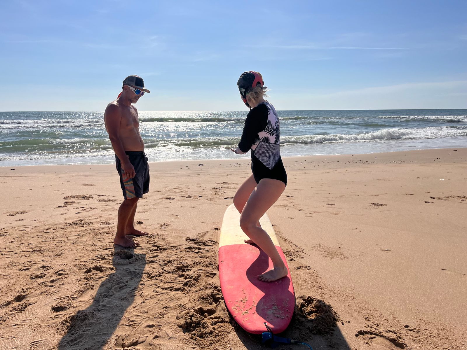 An instructor watches an adult beginner practise her stance on a surfboard on the sand.