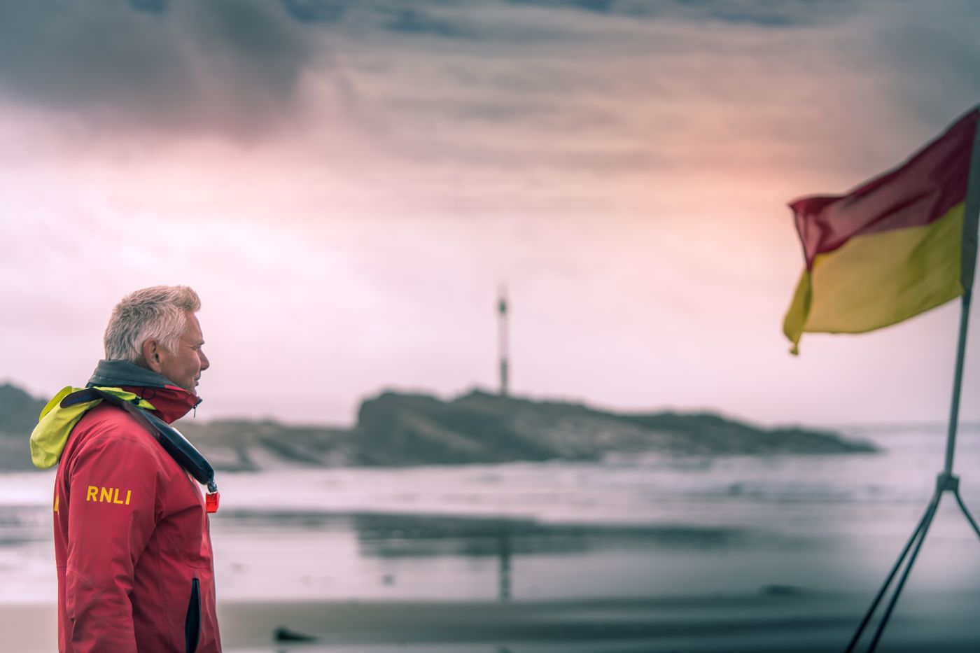 An RNLI lifeguard stands beside a red and yellow beach flag.
