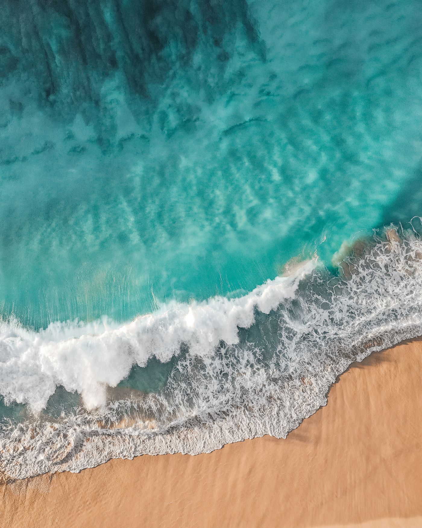 An aerial view shows successive white wave lines approaching a sandy shore.