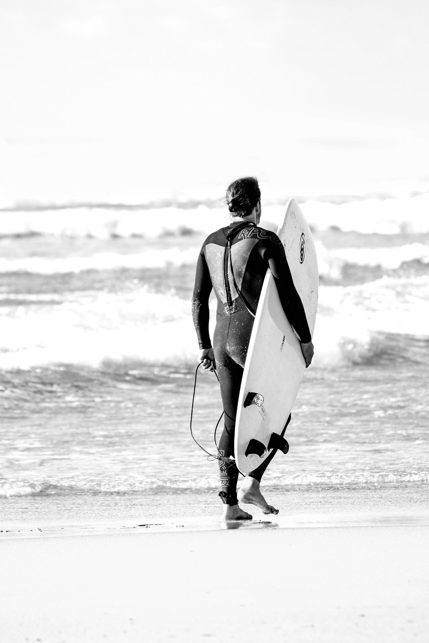 A surfer in a full wetsuit carries a board towards the shoreline.
