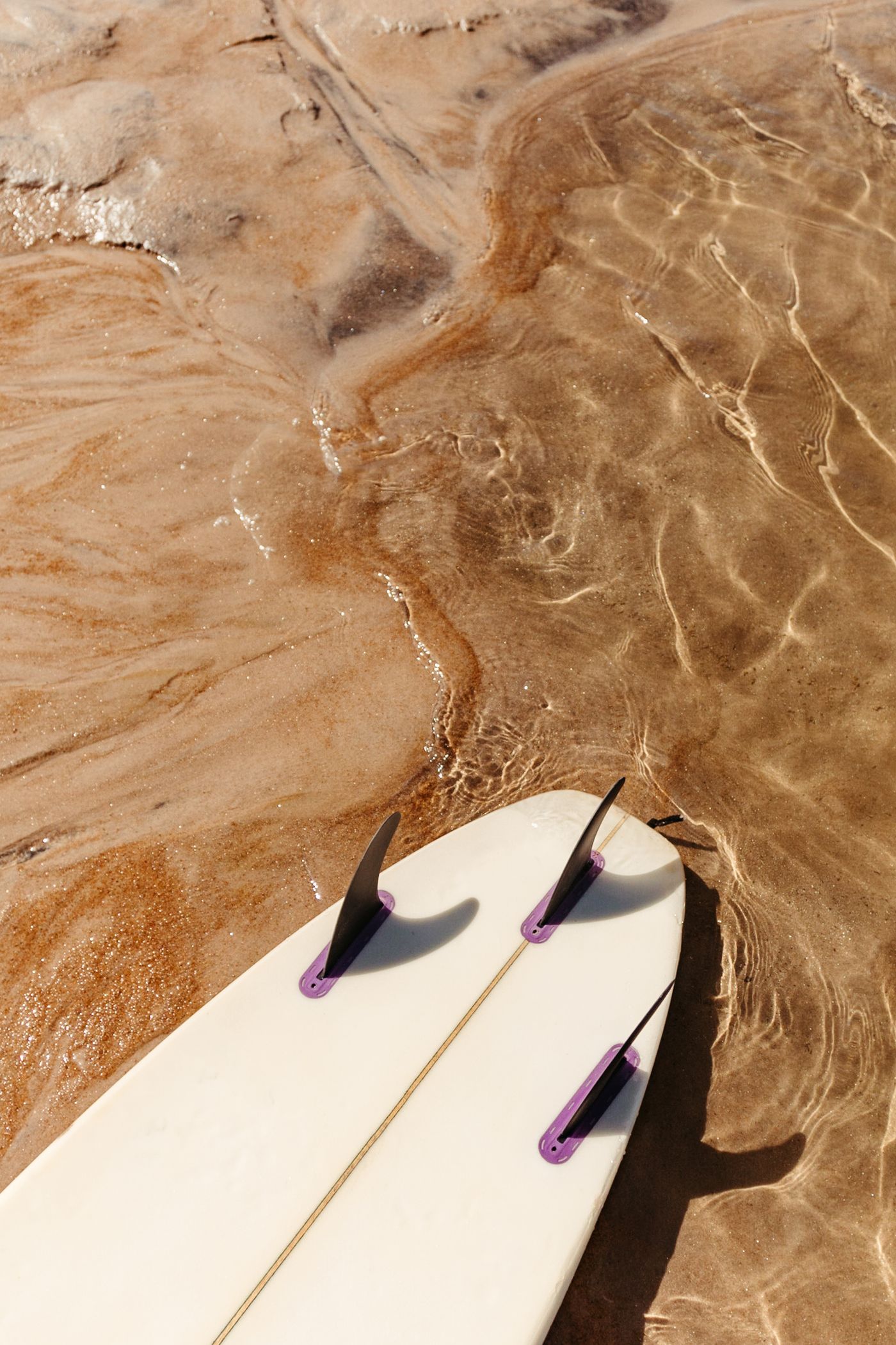 Three fins are visible on the underside of a surfboard lying beside shallow water.