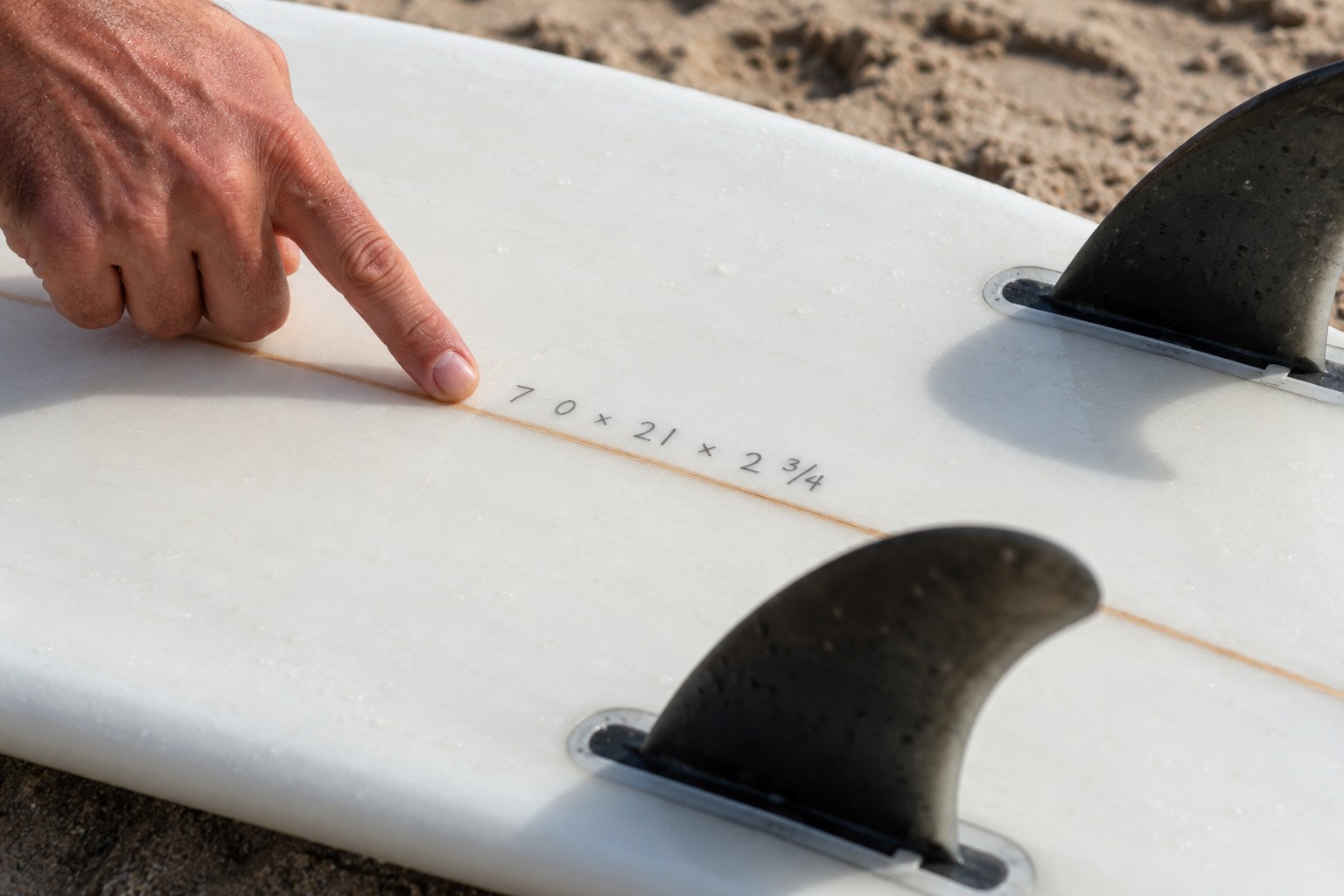 A hand points towards an illustrative dimensions line on the underside of a surfboard.