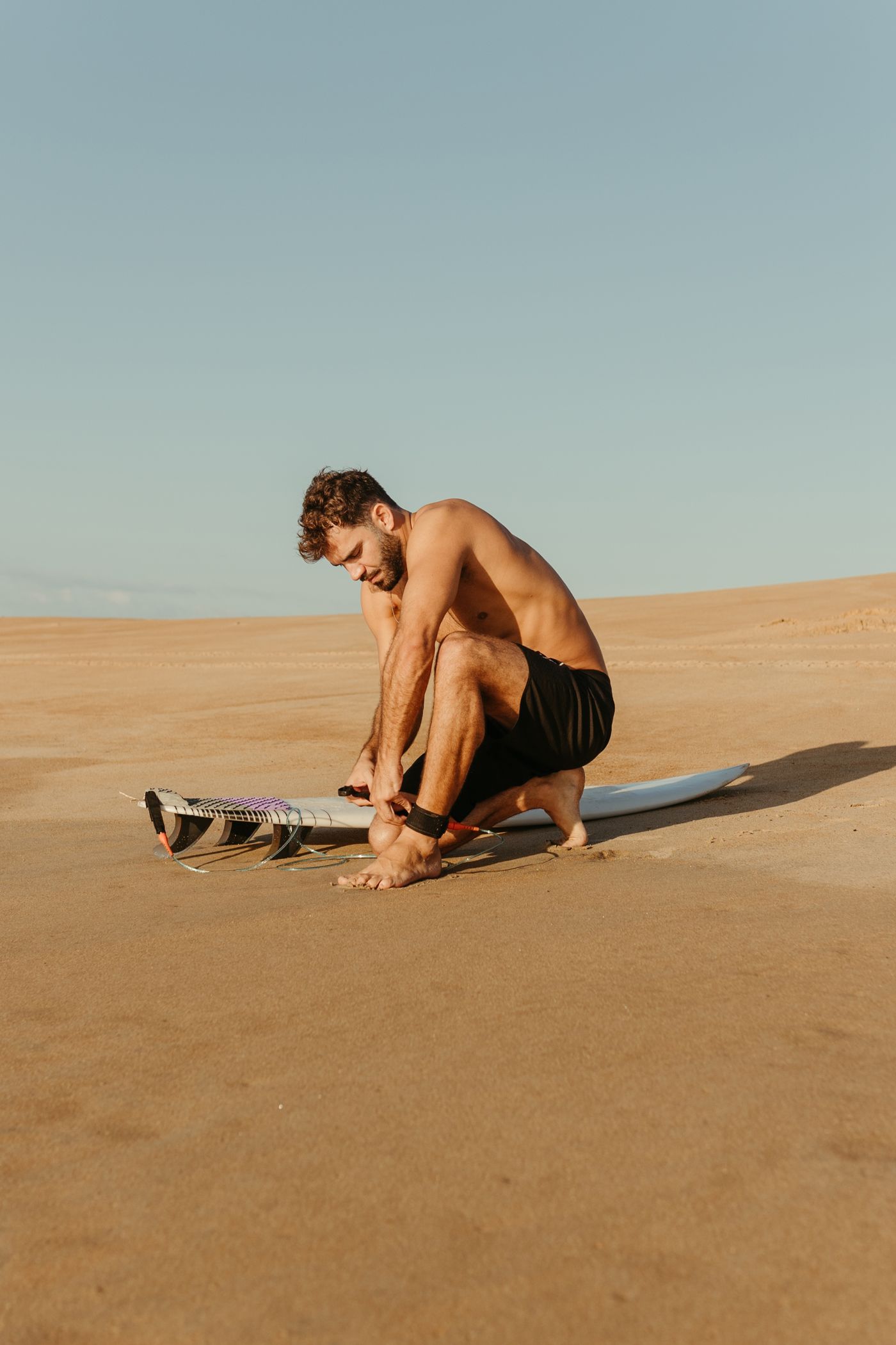 A surfer kneels on the sand to adjust the leash cuff at his ankle.
