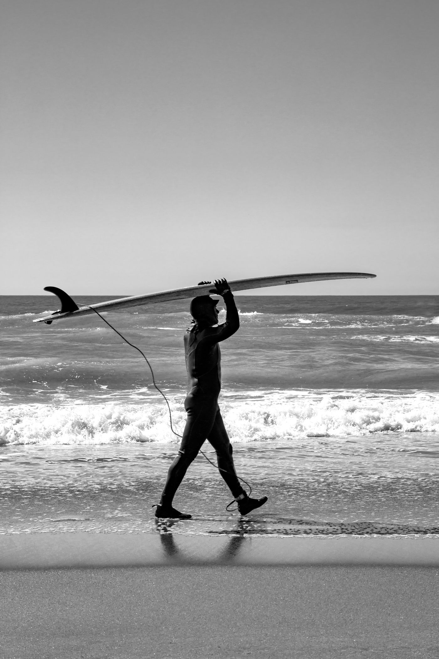 A surfer carries a long board overhead while walking along the shore.