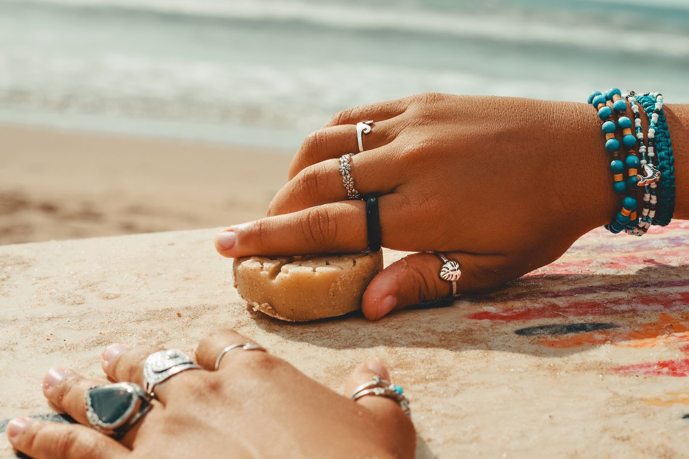 Hands rub a block of wax across a surfboard deck near the beach.