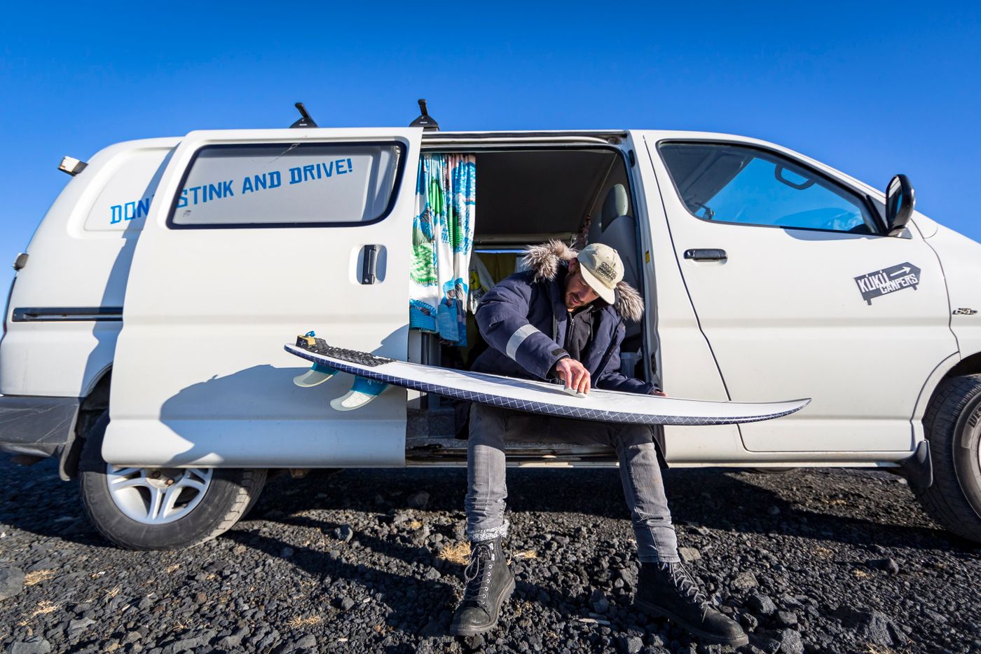 A surfer prepares a board beside the open door of a camper van.