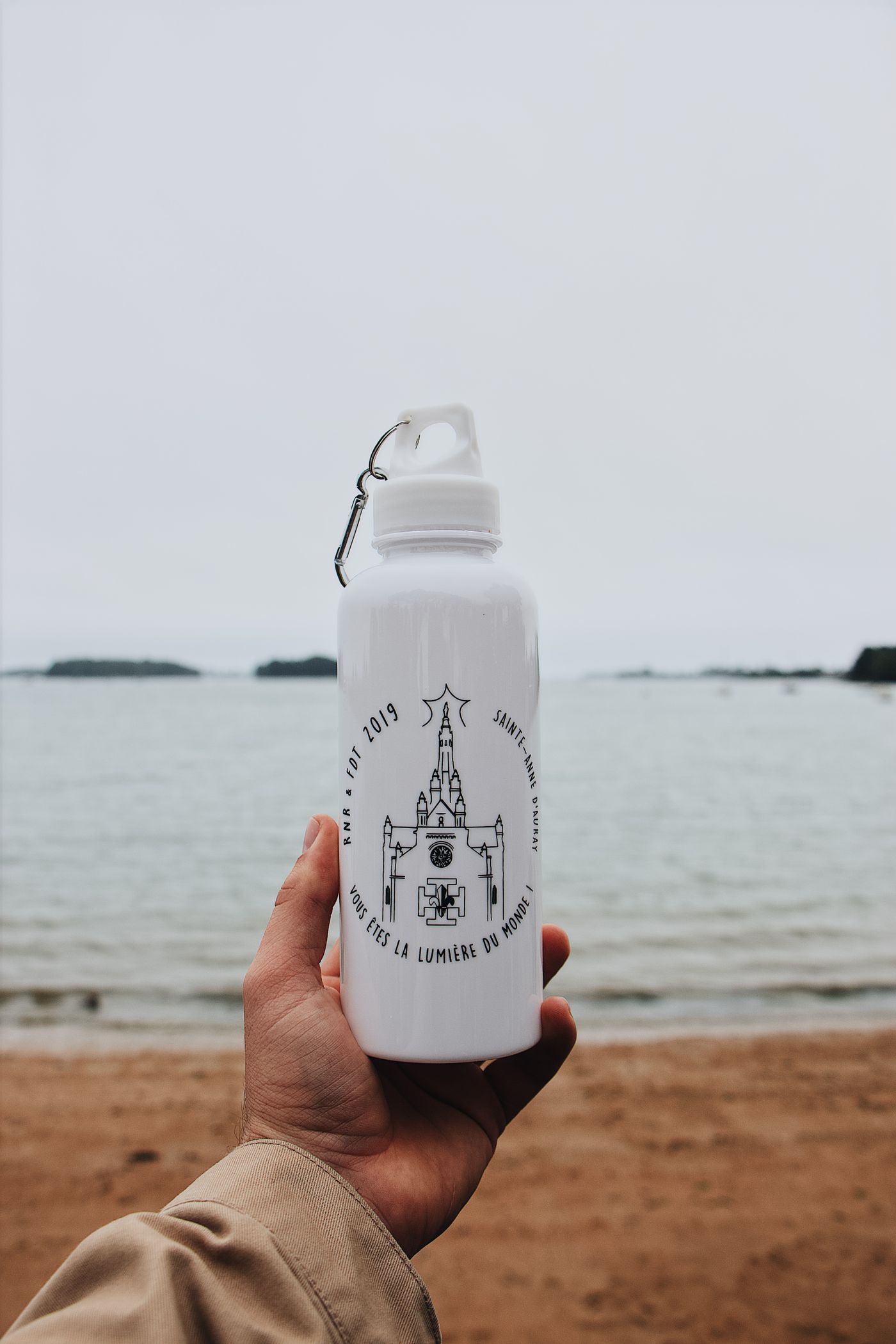 A hand holds a reusable white water bottle at the seaside.